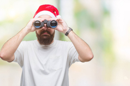 年轻的白种人时髦男人戴着圣诞帽，看着望远镜在孤立的背景上，聪明的脸上有自信的表情，严肃的思考