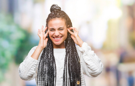 年轻的非洲裔美国女孩，梳着辫子，在孤立的背景下穿着毛衣，用手指捂着耳朵，因为嘈杂的音乐声而表情烦躁。聋子的概念。