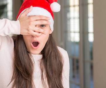 美丽的年轻女子戴着圣诞老人的帽子在家里，震惊地偷看着脸和眼睛，用手透过手指看着尴尬的表情。