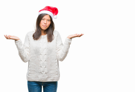 年轻的亚洲女人戴着圣诞帽，在孤立的背景上毫无头绪，表情混乱，举起手臂和手。 怀疑概念。