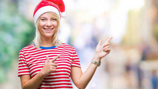 年轻漂亮的金发女人戴着圣诞帽，在孤立的背景上微笑着，看着镜头，双手和手指指向一边。
