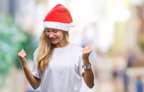 年轻漂亮的金发女人戴着圣诞帽，在孤立的背景下，非常高兴和兴奋地做着胜利者的手势，举起手臂，微笑着，尖叫着，争取成功。 庆祝概念。