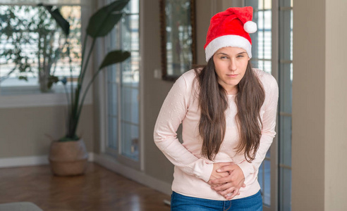 美丽的年轻女子戴着圣诞老人的帽子,在家里,手拿着肚子,因为消化不良