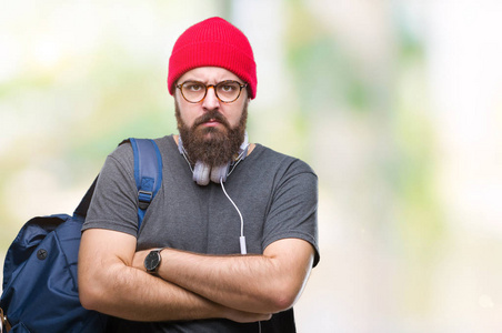 年轻的时髦男人戴着红色羊毛帽和背包，在孤立的背景怀疑和紧张，不赞成的表情在脸上交叉的手臂。消极的人。
