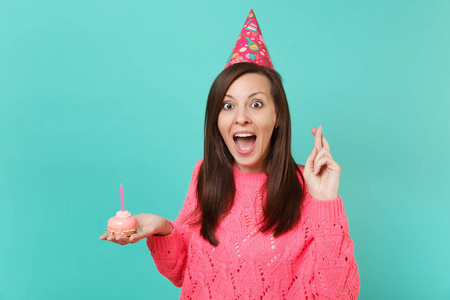 兴奋的年轻女孩穿着针织粉色毛衣生日帽，手指交叉，张开，手拿蛋糕，蓝色背景上的蜡烛隔离。人们的生活方式观念。把复制空间
