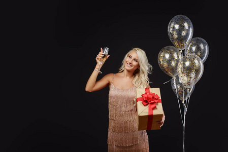快乐的年轻女子穿着豪华礼服，庆祝生日或圣诞派对，黑色背景上有纸屑，带着乐趣和微笑。 手拿礼物金气球和香槟