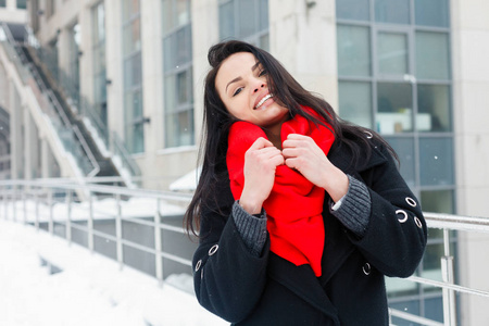 冬季圣诞节的生活方式，假日和时尚概念，美丽的年轻微笑的女人，穿着黑色外套和红领巾在冬季城市摆姿势。 下雪
