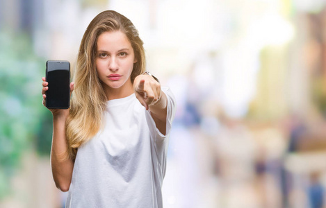 年轻漂亮的金发女人在孤立的背景上显示智能手机屏幕，手指指向相机，并向你手势正面和自信的手势。