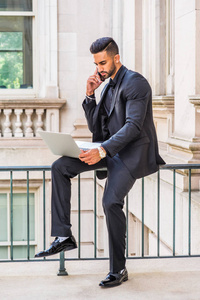 年轻的东印度裔美国商人，留着胡子，在纽约工作，穿着黑色西装，皮鞋，坐在办公楼里的栏杆上，工作着笔记本电脑，讲着手机