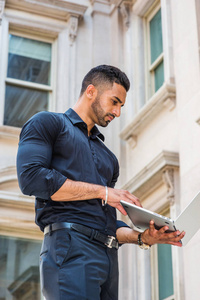 通往成功的道路。 年轻的东印度美国商人，留着胡子，在纽约工作，穿着黑色衬衫，手提电脑，站在老式办公楼外面看书