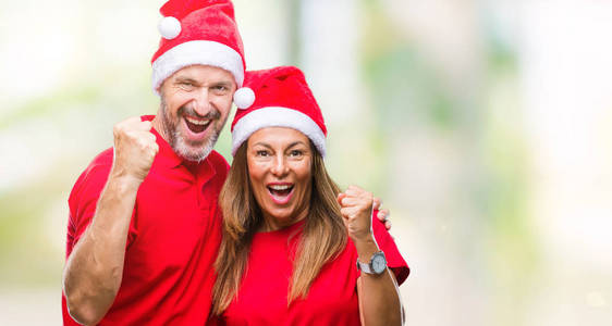 中年西班牙裔夫妇戴着圣诞帽，在孤立的背景上庆祝，惊讶和惊讶的成功，举起手臂和睁开眼睛。 赢家概念。
