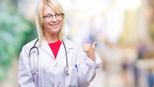 年轻漂亮的金发医生女人穿着医疗制服，在孤立的背景上微笑，快乐的脸，看着，用拇指指向一边。