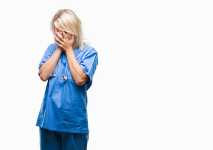 年轻漂亮的金发医生女人穿着医疗制服，在孤立的背景上，悲伤的表情覆盖着脸，双手哭泣。 抑郁的概念。