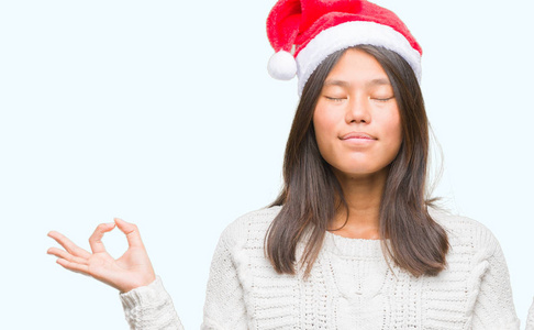 年轻的亚洲女人戴着圣诞帽，在孤立的背景上放松，闭着眼睛微笑，用手指做冥想手势。 瑜伽概念。
