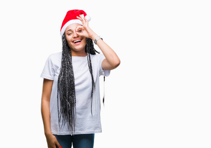 年轻的辫子非洲裔美国女孩戴着圣诞帽，在孤立的背景下做着好的手势，用手微笑，用眼睛透过手指看着快乐的脸。