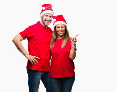 中年西班牙裔夫妇戴着圣诞帽在孤立的背景上，脸上带着大大的微笑，用手和手指指向一边，看着相机。