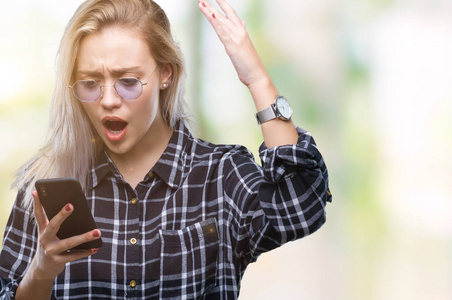 年轻的金发女人用智能手机在孤立的背景下发短信，愤怒和沮丧地大喊大叫，疯狂地举起手大喊大叫，愤怒的概念