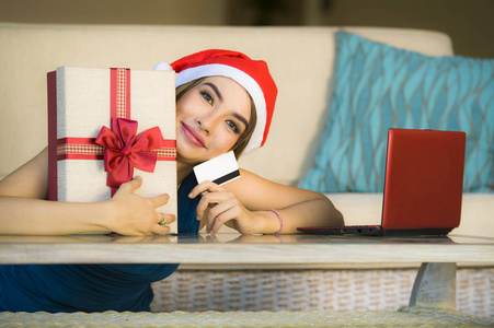 年轻快乐和美丽的女孩在家中放松，沙发上戴着圣诞帽，用笔记本电脑支付圣诞礼物，信用卡在网上购物和网络商务中微笑