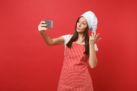 家庭主妇女厨师或面包师穿条纹围裙，白色T恤，烤厨师帽子隔离在红墙背景上。 微笑有趣的女人在手机上做自拍。 模拟复制空间概念