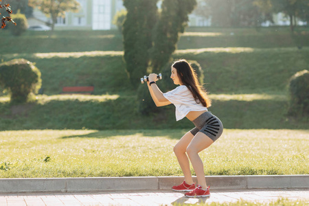 舒适的健身女孩在日落时在外面的草地上用哑铃做运动