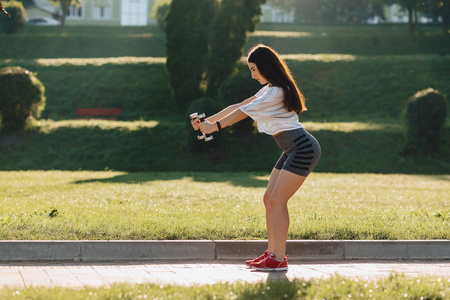 舒适的健身女孩在日落时在外面的草地上用哑铃做运动