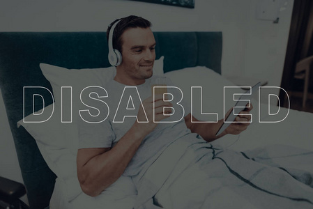 残疾人。 男人躺在床上。 男人在平板电脑上看视频。 戴着耳机的人。 拿着果汁的杯子的人。 男人在微笑。 男人位于现代卧室。 轮椅