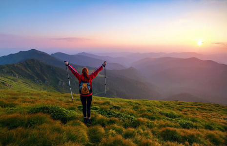 红头发的女运动员带着背包和棍子站在绿色的山丘上，看着高山景观和迷人的日落。