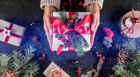 女性手拿着圣诞礼品盒，上面有红色丝带，节日背景是杉树枝松果。 圣诞节和新年快乐卡波克闪闪发光。 平躺式顶部视图