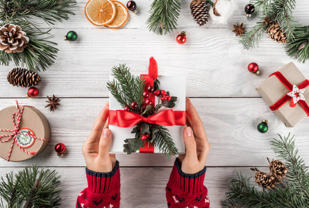 女性手拿着圣诞礼品盒在白色的木制背景上与杉树枝松果。 圣诞节和新年快乐主题博凯赫闪闪发光。 平躺顶部视图。