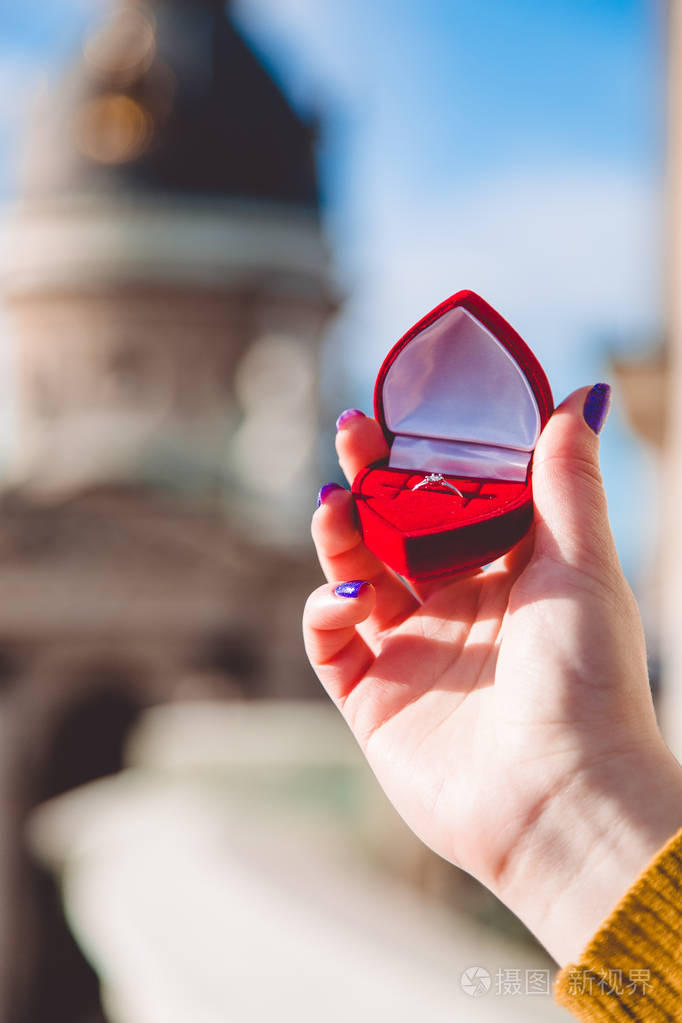 女人的手拿着订婚戒指戴着钻石在建筑模糊背景前的红色心形盒子里求婚