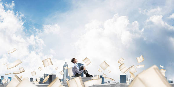 商人一边看一边坐在一堆文件上，一边在背景上云云般的天空中飞舞的书中。 混合媒体。