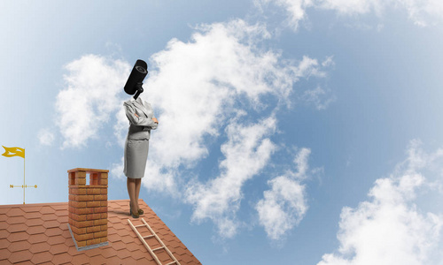 没有脸的女商人用相机变焦而不是头站在房子的屋顶上