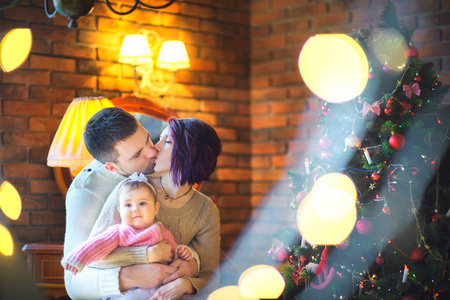 幸福的家庭在明亮的灯光下坐在节日的圣诞树旁拥抱和亲吻