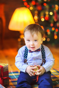 有趣的婴儿坐在地板上，手里拿着一件小礼物，在明亮的节日灯光的背景下微笑。