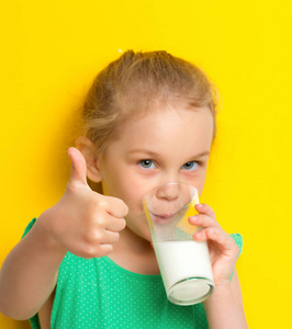 可爱的蓝眼睛小女孩正在用玻璃烧杯喝牛奶，在黄色的背景上竖起大拇指上课很好吃。