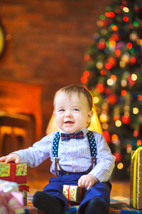 有趣的婴儿坐在地板上，手里拿着一件小礼物，在明亮的节日灯光的背景下微笑。