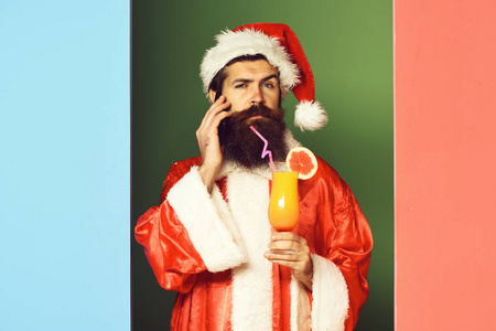 帅有胡子的圣诞老人人与非酒精性鸡尾酒杯举行圣诞节或圣诞毛衣和新年帽子上多彩的演播室背景严重脸上长长的胡须