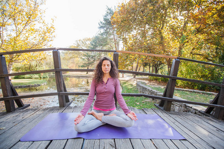 女人冥想和练习瑜伽莲花姿势帕德马萨纳。 在阳光明媚的秋日在城市公园的木桥上冥想。