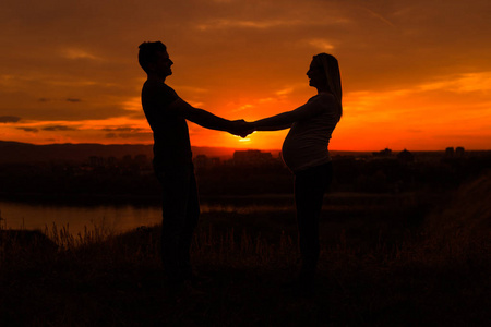 丈夫和怀孕的妻子的剪影，牵着手，享受在一起的户外时光。色调的形象。