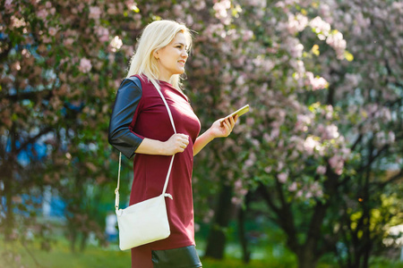 在春暖花开的公园里使用智能手机的年轻金发女人的肖像。