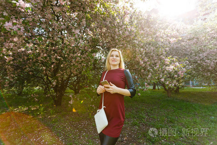 在春暖花开的公园里使用智能手机的年轻金发女人的肖像。