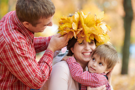 父子把黄色的落叶放在母亲的头上。 幸福的家庭在秋天的城市公园。 孩子和父母。 他们摆出微笑的姿势，玩得很开心。 明亮的黄色树木。