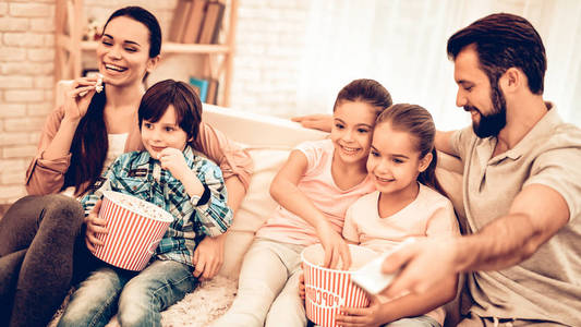 可爱开朗的家人在家看电影。 孩子们在家坐在沙发上吃爆米花和微笑。 小可爱的孩子带着玩具，父母在沙发上拥抱看动画片