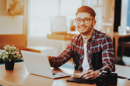 微笑自由职业者在笔记本电脑上工作的肖像。 时髦的平面设计师戴着眼镜，坐在桌子上，用数码平板电脑在室内用笔触摸照片。 远程工作概念