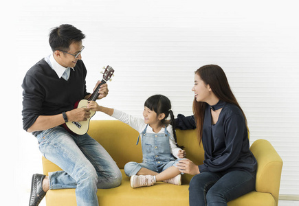 亚洲女儿弹吉他，和爸爸妈妈在客厅的沙发上唱歌。 概念家庭享受。