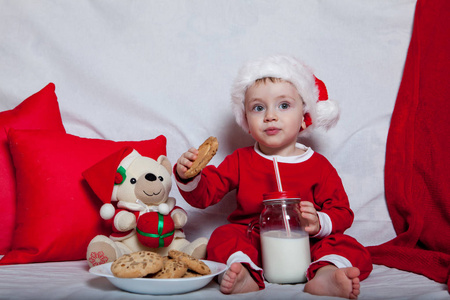 一个戴着红帽子的孩子吃饼干和牛奶。 戴着红色帽子的婴儿的圣诞摄影。 新年假期和圣诞节。
