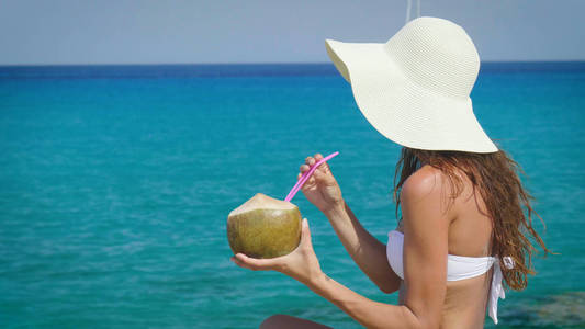 美丽的年轻女孩在海边喝椰子鸡尾酒，戴着白色比基尼太阳镜，金色的沙子背景。 概念海上度假自由阳光旅行海浪新鲜水果度假饮食热。