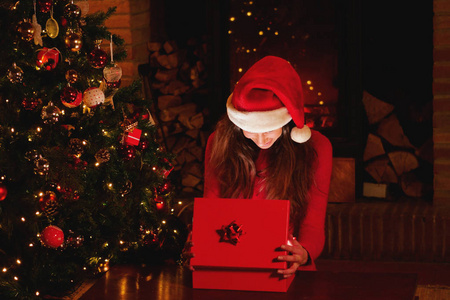 在圣诞节，一个美丽的女人在圣诞树下打开她的礼物，当她打开礼物时，她像魔法一样亮起。 概念魔术圣诞礼物假期。