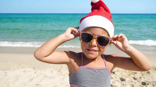 一个小女孩正在海上玩，戴着圣诞老人的帽子，穿着泳衣，戴着太阳镜，背景是深蓝色的海水。