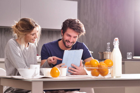 厨房里的一对夫妇看着平板电脑，一边吃早餐，一边开心地笑着，一边看着他们假期或过去时光的纪念品照片。 概念家庭技术记忆。
